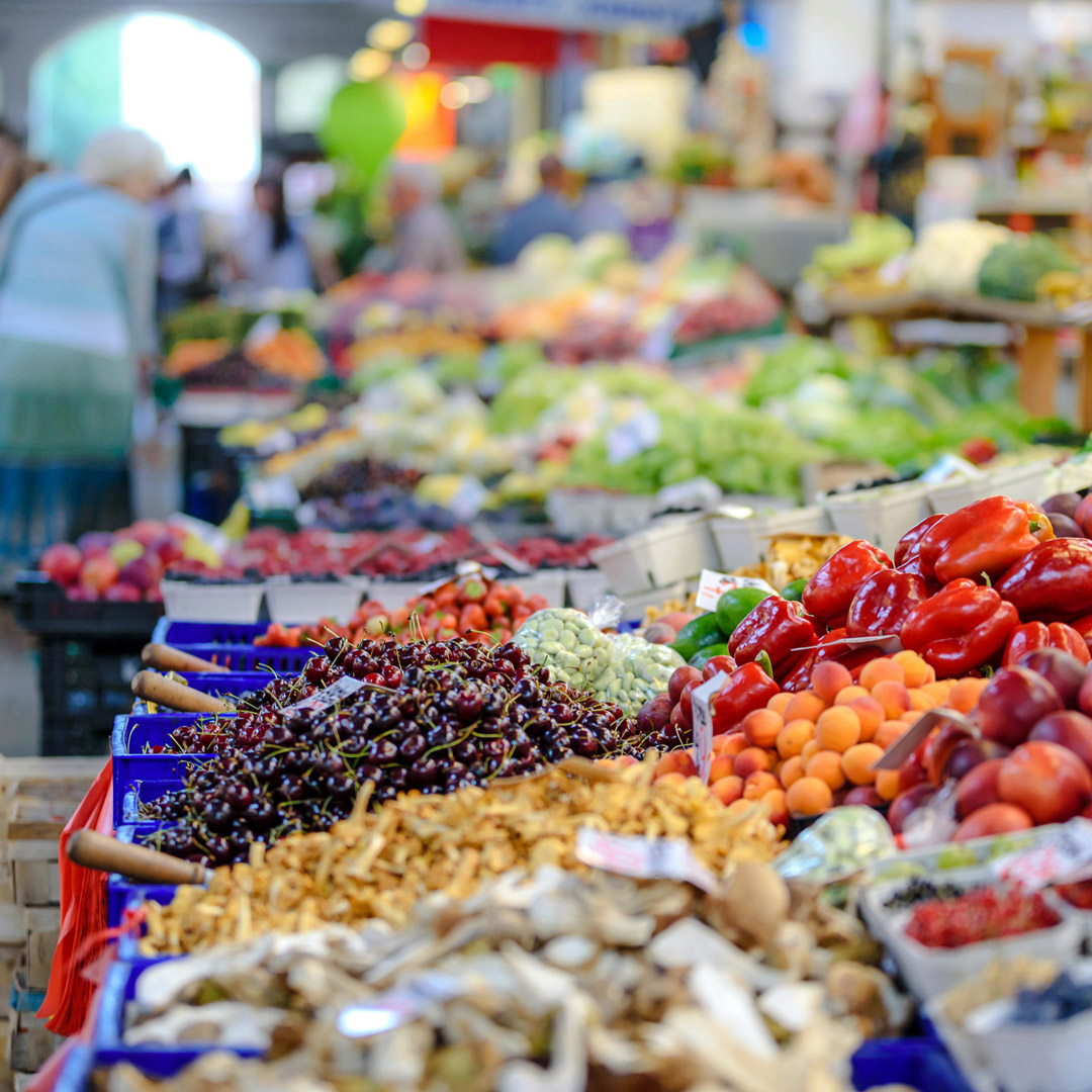 Una parada de fruites i verdures en un mercat local. Es veuen pebrots vermells, cireres i préssecs. Al fons, desenfocat, es poden veure alguns clients i venedors